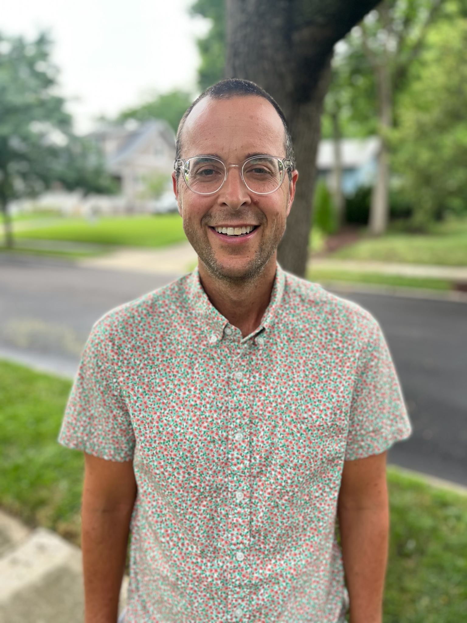 Smiling man wearing glasses and a floral shirt, standing outside on a neighborhood street